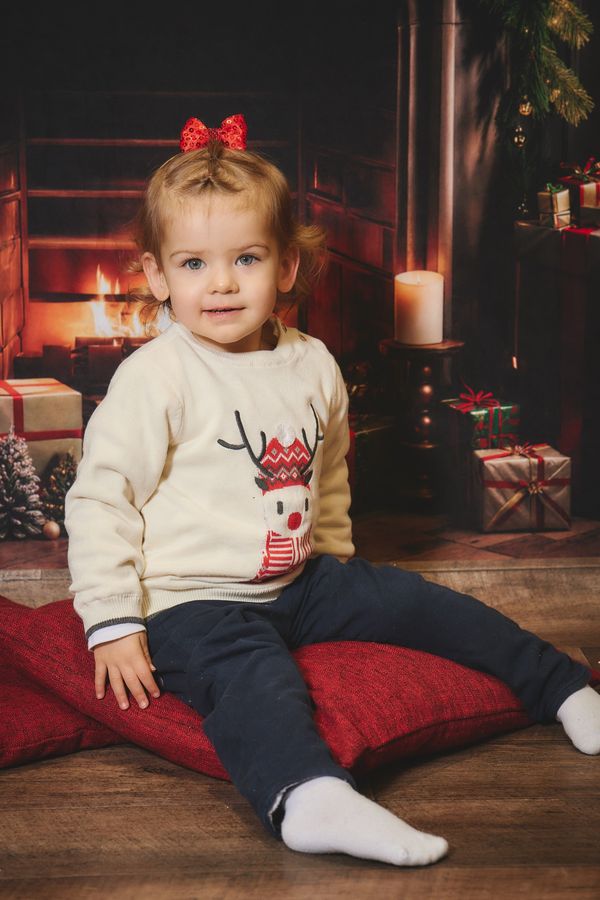 Petite fille au nœud rouge et pull à renne assise sur un coussin rouge, décor de Noël avec cheminée, chez Aubert à Coudekerque-Branche