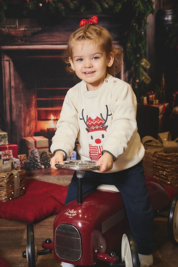 Petite fille souriante sur une voiture porteur rouge à l'ancienne, devant une cheminée de Noël, chez Aubert