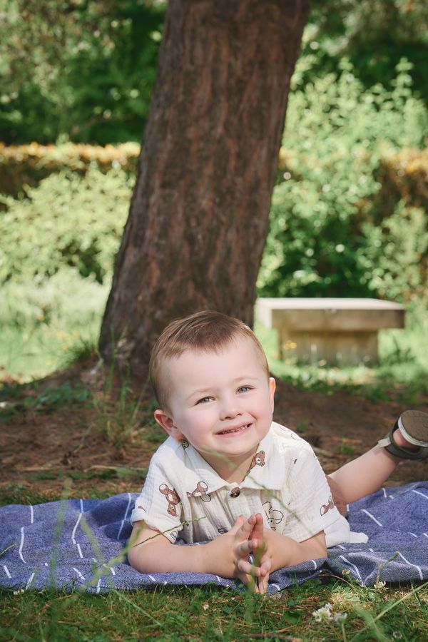 Petit garçon de 3 ans allongé sur un plaid bleu, mains jointes et grand sourire, sous un pin des Jardins du Colysée